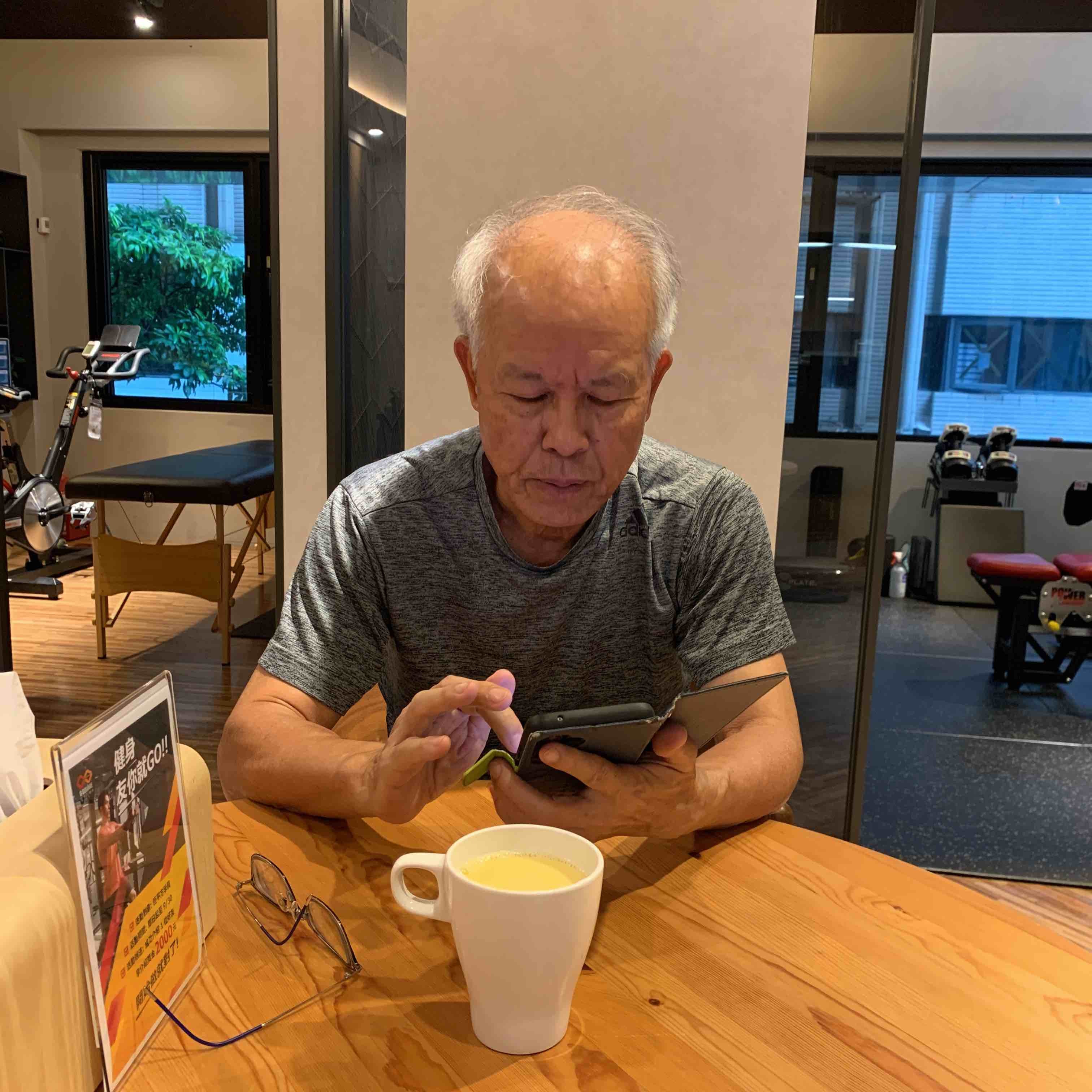 張立秋坐在桌前使用手機，桌上有杯子。